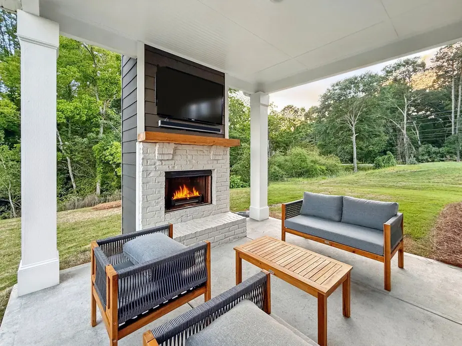 Covered patio at dusk with a lit stone fireplace, a wall-mounted TV over woven rope furniture, and a wooded backyard beyond